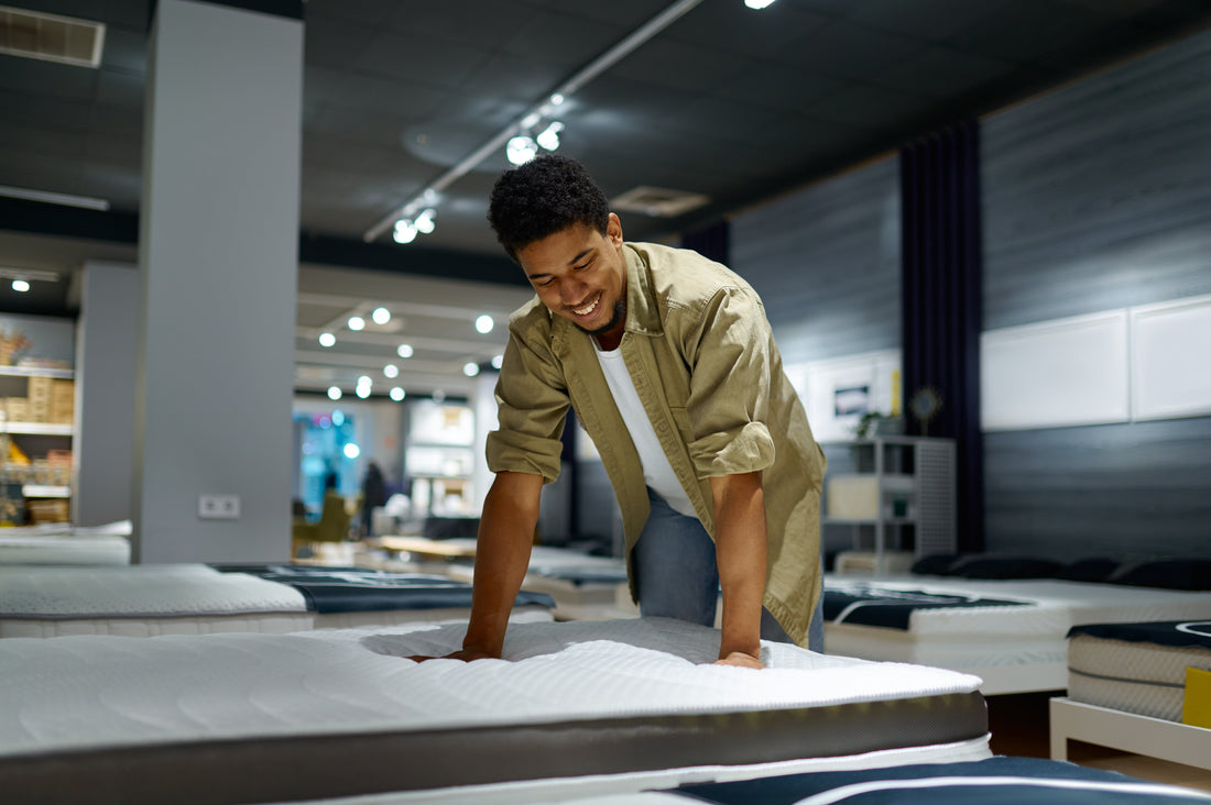 A man in a mattress store smiling at a mattress and pressing firmly with both hands onto the mattress.