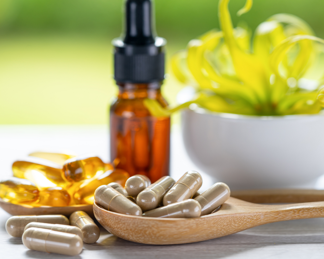 Daily vitamins in capsule format in a wooden spoon with a liquid vitamin dropper bottle and small plant in the background.